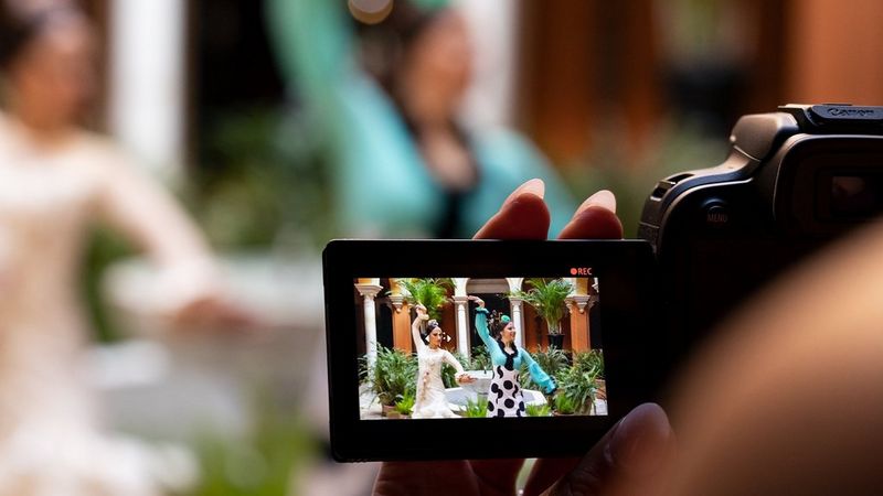 A woman’s hand holding the LCD screen of EOS R10 showing the camera taking a video of flamenco dancers visible in the background.