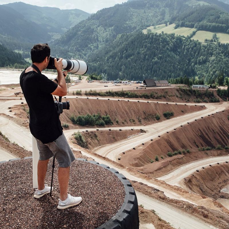 A man stands on a raised platform above a sandy racetrack holding a Canon EOS R3 with a super-telephoto lens attached. 