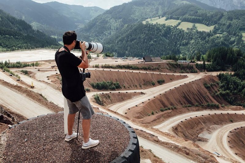 A man stands on a raised platform above a sandy racetrack holding a Canon EOS R3 with a super-telephoto lens attached.