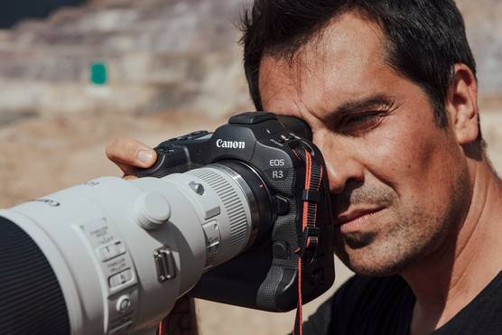 A male photographer looks through the viewfinder of a Canon EOS R3 with a long lens attached.