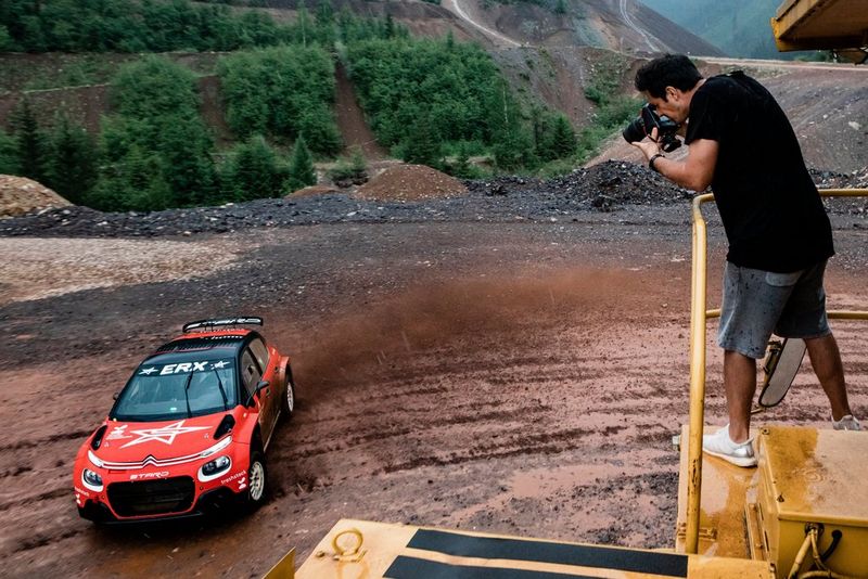 A man stands on an elevated gantry with a camera raised to his eye, pointing it down at a red rally car on the ground.