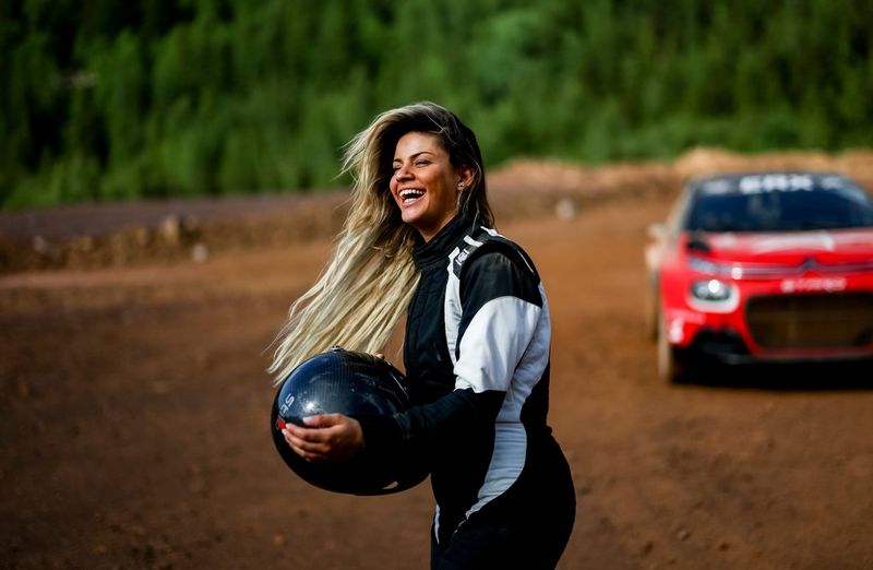 A woman in a black and white racing suit stands holding her helmet, long hair blowing in the wind. A red rally car is behind her, out of focus.
