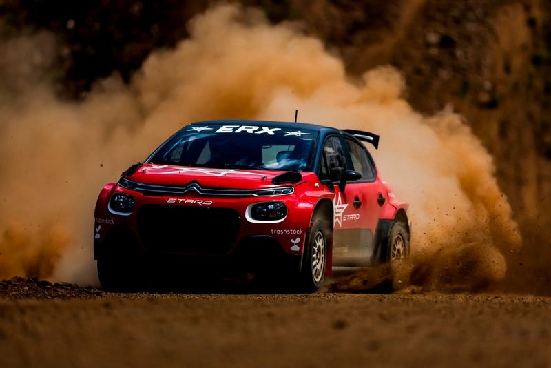 A red rally car captured at speed, kicking up a cloud of dust behind it. 