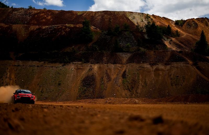 A red rally car drives towards the camera, entering the shot from the left in front of a steep hill.