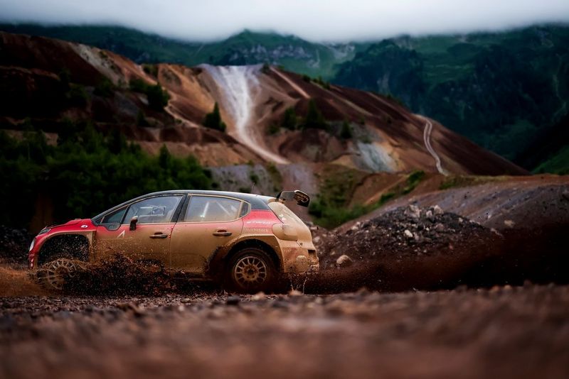 A dirty red rally car races across the image, kicking up a cloud of dust behind it, in front of an out-of-focus mountainous background.