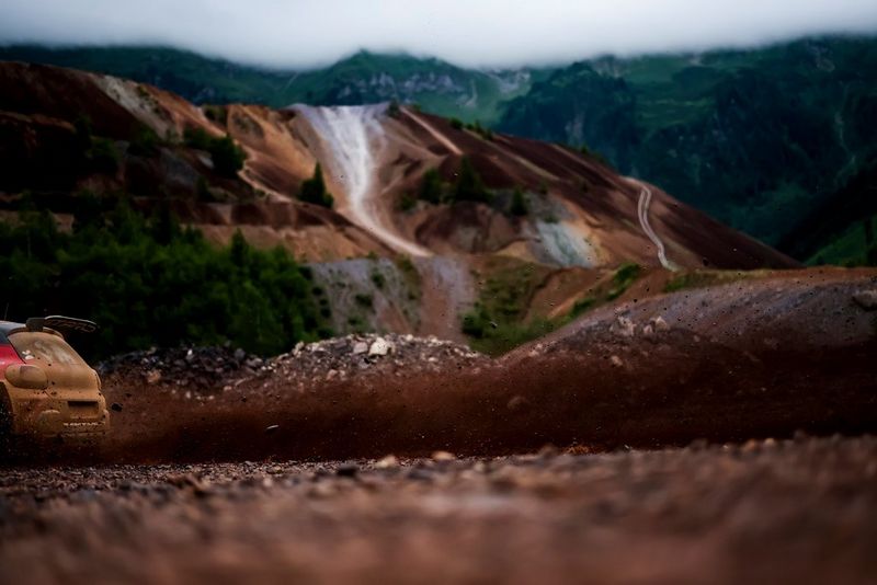 The back of a red rally car is seen at the very left of the shot, exiting the frame. The car is still in focus, and the mountain background to the picture is blurred.