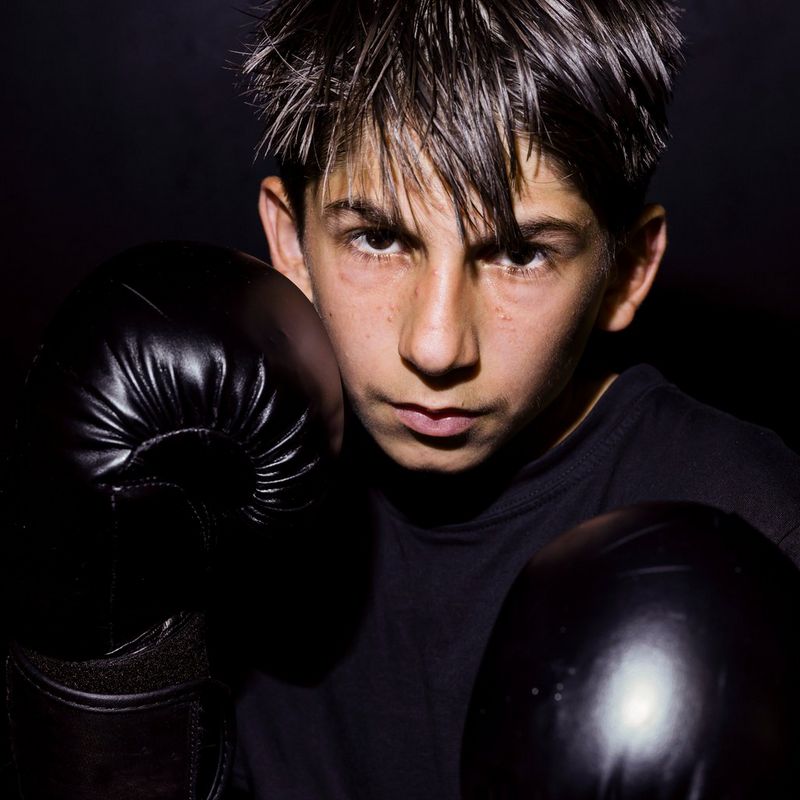 Un ragazzo con i capelli scuri, che indossa guanti da kick boxing, in piedi contro uno sfondo scuro, con i pugni alzati davanti al viso e al torace, in posizione di combattimento.