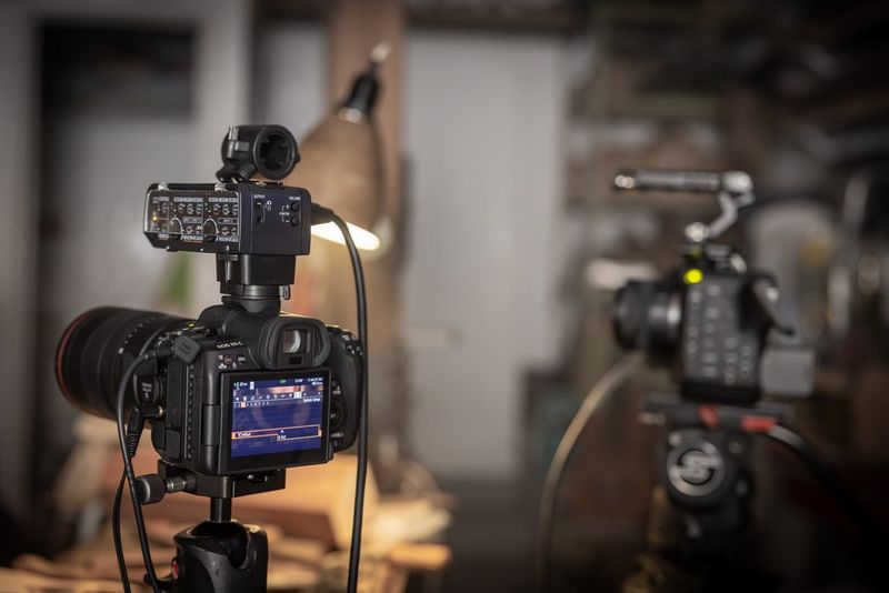 The rear of a Canon EOS R5 C hybrid camera, mounted on a tripod, showing the bottom half of a man's face in the LCD touchscreen. 