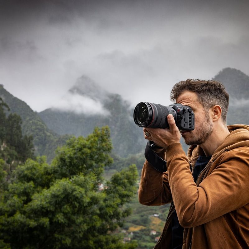 A man in an orange hooded jacket holding a Canon EOS R5 C camera in front of a backdrop of cloud-covered mountains.