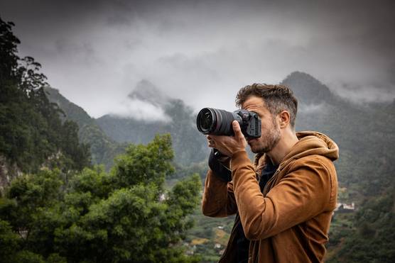  A man in an orange hooded jacket holding a Canon 中国福彩网5 C camera in front of a backdrop of cloud-covered mountains.