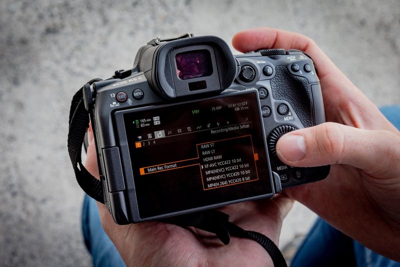 A pair of hands hold a Canon EOS R5 C camera, navigating the Main Recording Format screen. 