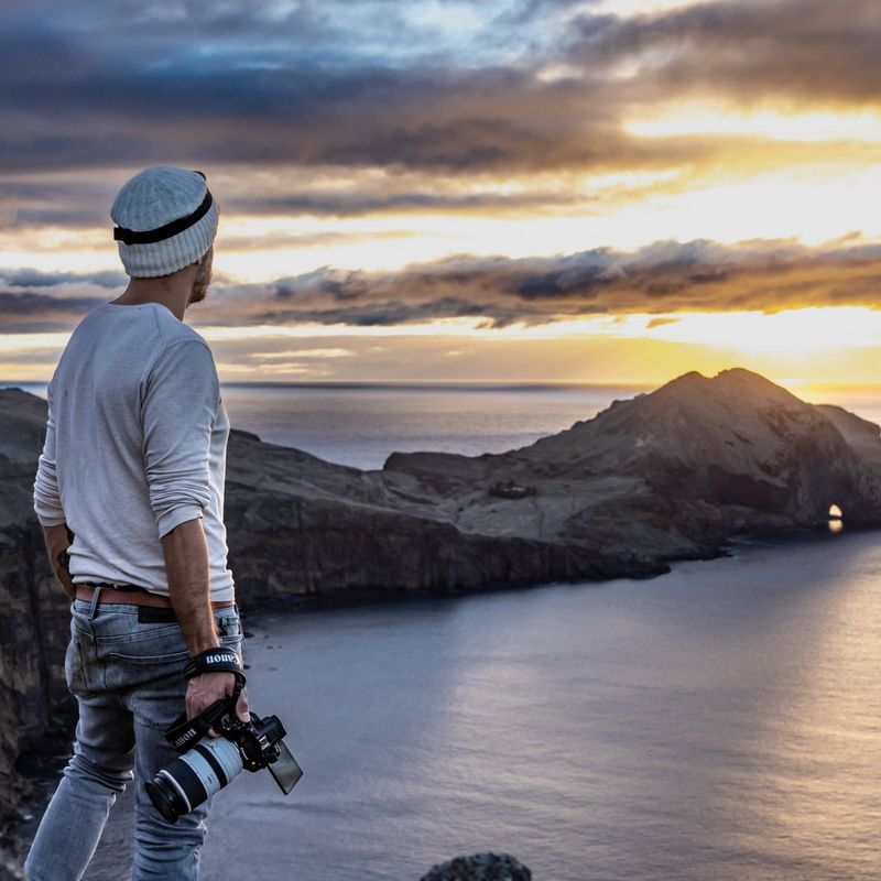 A man wearing a winter hat and holding a Canon EOS R5 C looks out over a bay as the sun breaks through the clouds.