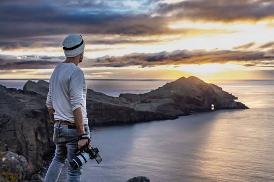 A man wearing a winter hat and holding a Canon 新万博体育_新万博体育官网- 【长期稳定】@5 C looks out over a bay as the sun breaks through the clouds. 