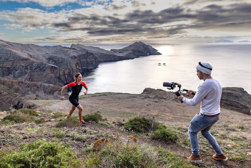 Using a Canon EOS R5 C video camera, a videographer films a runner on a clifftop, with the rocky shoreline and dark clouds in the background.