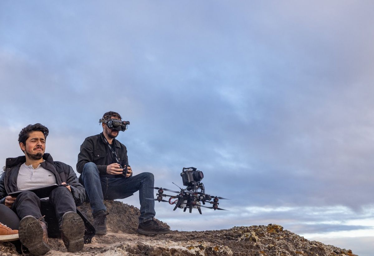 Two men sit together at the edge of a cliff. The man on the right is wearing goggles and holding a controller while a Canon EOS R5 C attached to the top of an FPV drone flies nearby.