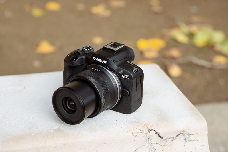 Камера Canon EOS R50 с комплектным объективом в разложенном состоянии лежит на белой поверхности.