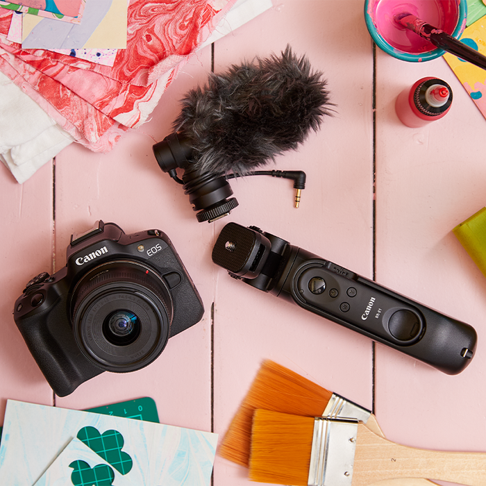 A flatlay of a Canon EOS R50 camera, external microphone and tripod grip on a pink wooden background, surrounded by crafting materials.