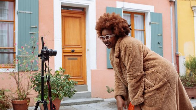 Social media influencer Fatou N'Diaye, wearing a fluffy brown coat, crouches and smiles in front of a Canon EOS R50 camera on a tripod.