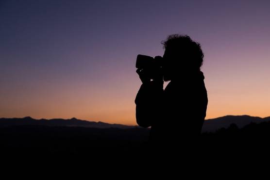A man faces sideways, holding a camera to his face, silhouetted against a purple and orange sky at sunset.