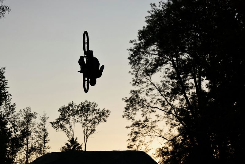 A mountain biker mid-stunt, silhouetted against the sky and framed by trees.