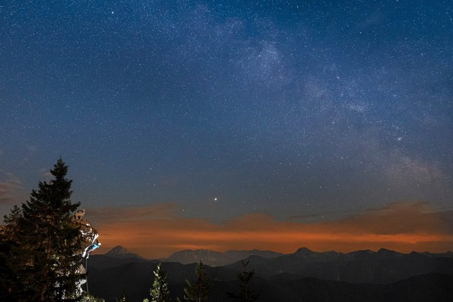 Um céu estrelado, o horizonte com faixas laranja e um abeto apenas visível no primeiro plano à esquerda.
