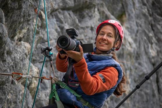 Ulla Lohmann holds a Canon EOS R5 camera as she hangs from a sheer rock face.