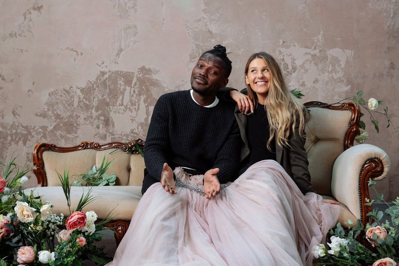 Il presentatore e creatore di contenuti Tomi Adebayo e la fotografa Rosie Hardy si siedono insieme su una chaise longue, un abito da ballo rosa chiaro sistemato sulle ginocchia e fiori su entrambi i lati.
