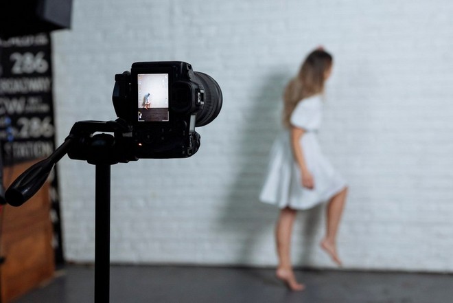 La fotografa Rosie Hardy è in piedi su una gamba sola, indossa un abito bianco, contro un muro bianco, con una Canon EOS R5 montata su un treppiede davanti a lei.