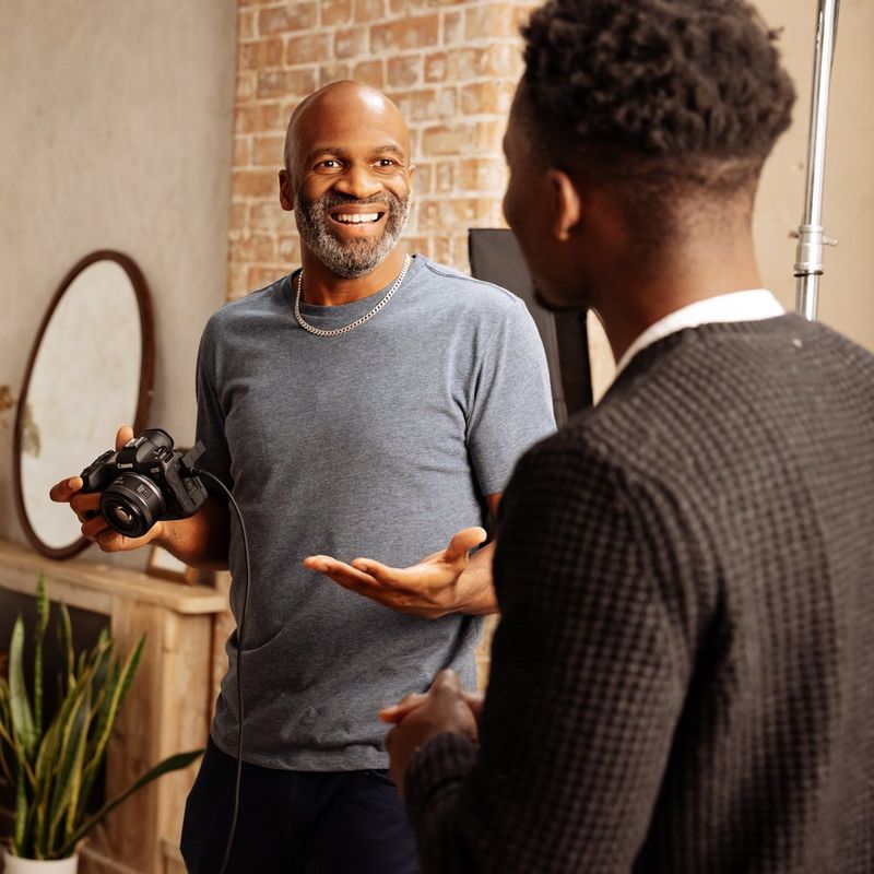 A man holding a Canon camera smiles and talks to another man.