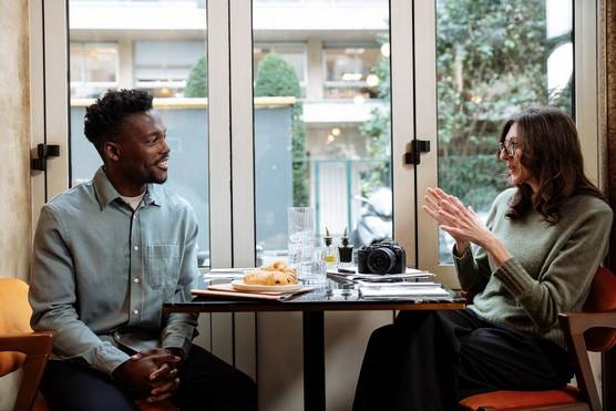 Presenter Tomi Adebayo and content creator Laura Hannoun sit at a table next to a window in a Parisian cafe.