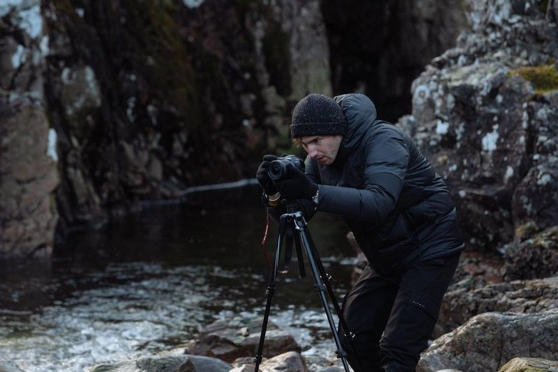 Un fotografo in abiti invernali, accovacciato dietro una fotocamera Canon EOS R6 montata su un treppiede posizionato sulle rocce lungo un fiume.