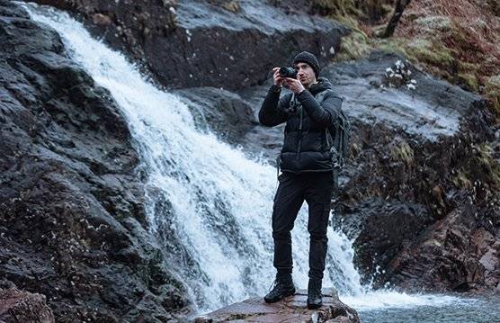 Videographer and photographer Harrison Brown holds a Canon camera, with a waterfall flowing down the rocks behind him.