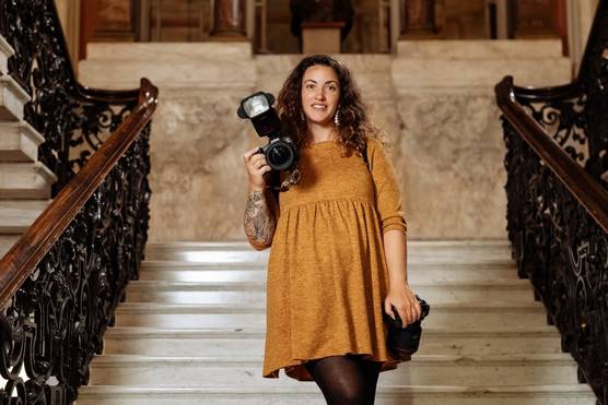 Photographer Alison Bounce stands in the stairway of a grand building, holding up a Canon EOS R5 camera while another hangs from a harness at her side.