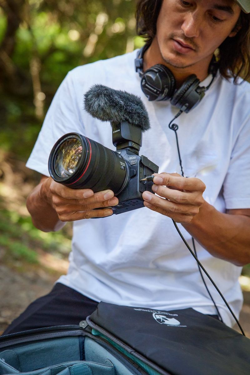 A man with headphones around his neck sits on the ground, plugging them into a Canon EOS R6 Mark II, which also has a microphone attached on top.