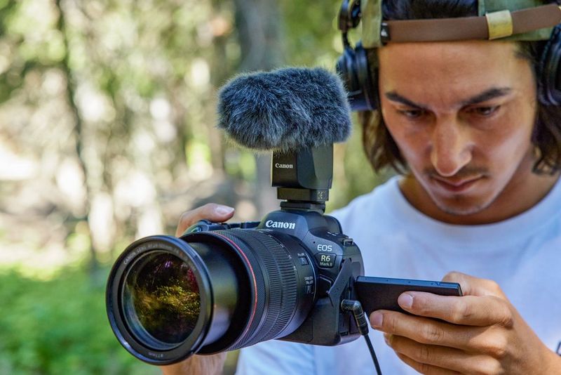 A man wearing over-ear headphones holds a Canon EOS R6 Mark II camera with a telephoto lens and microphone attached, using the folded-out touchscreen.