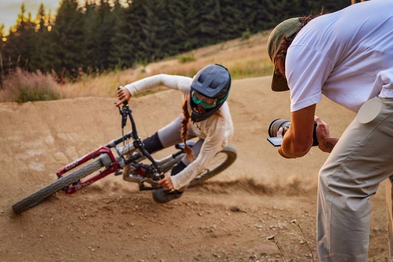 A man leans down holding a Canon EOS R6 Mark II to video a woman cycling around a tight curve.