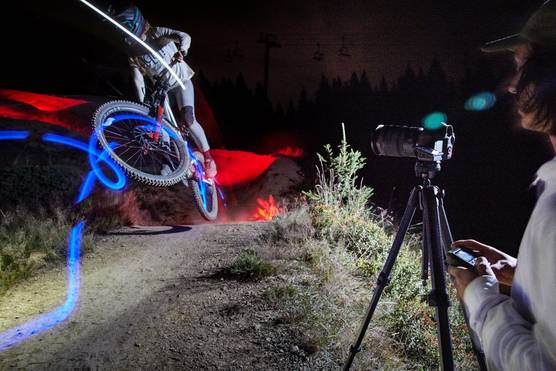 A mountain biker surrounded by red and blue light trails performs a jump against a dark sky. A Canon 新万博体育_新万博体育官网- 【长期稳定】@6 Mark II is on a tripod at the side of the dirt track.