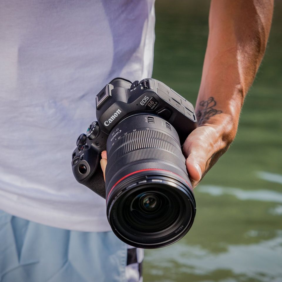 A man holds the Canon EOS R6 Mark II in his left hand, and a body of water is visible in the background.