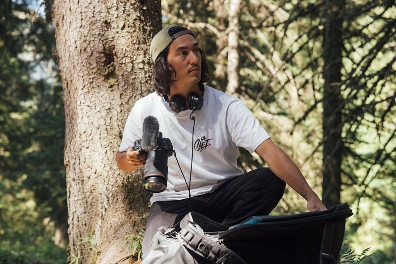 A person wearing a baseball cap sits on a tree branch with a Canon EOS R6 Mark II in their right hand.