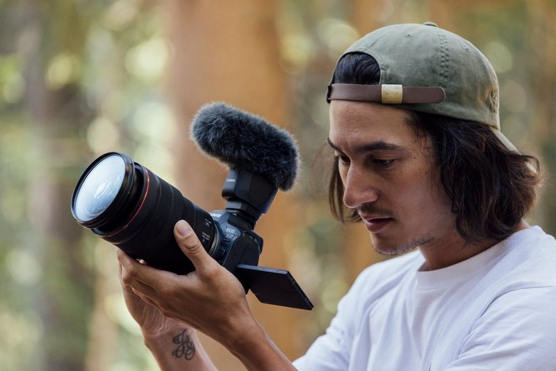 A person wearing a baseball cap looks at the screen of a Canon EOS R6 Mark II camera in a forest setting.