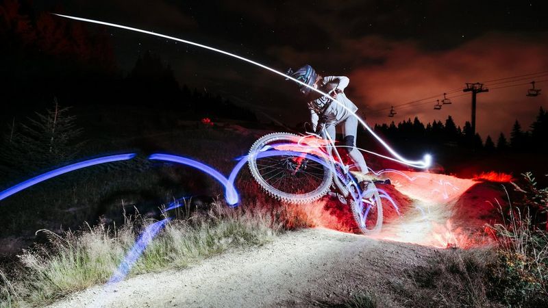 Sur une photo prise avec un Canon EOS R6 Mark II, les roues d'un vététiste quittent le sol alors que ce dernier roule dans l'obscurité, entouré de traînées lumineuses colorées.