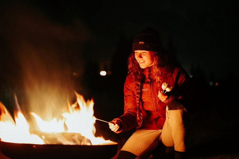 Una donna in abiti invernali sorride mentre arrostisce un marshmallow su un bastoncino su un falò di notte. Scattata con Canon EOS R6 Mark II.