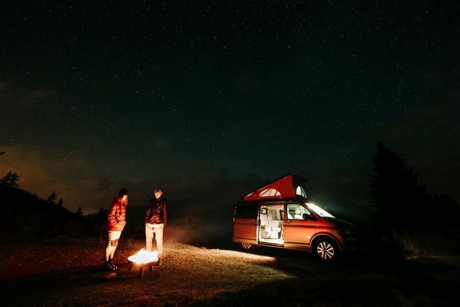 A photo taken on a Canon EOS R6 Mark II of two people standing in the dark, lit only by a small fire in front of them and the light from the open door of a van beside them.