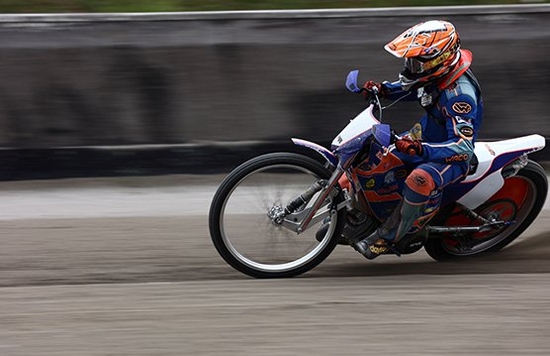 A person riding a dirt bike around a track, leaning the bike low to the ground, photographed using camera panning. © Richard Walch