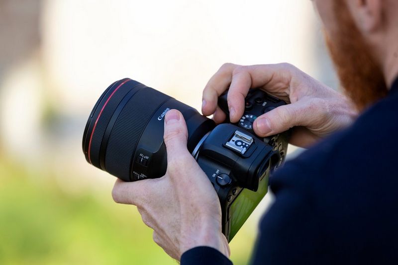 Мужчина держит в руках камеру Canon с объективом и одной рукой регулирует положение диска установки режима.