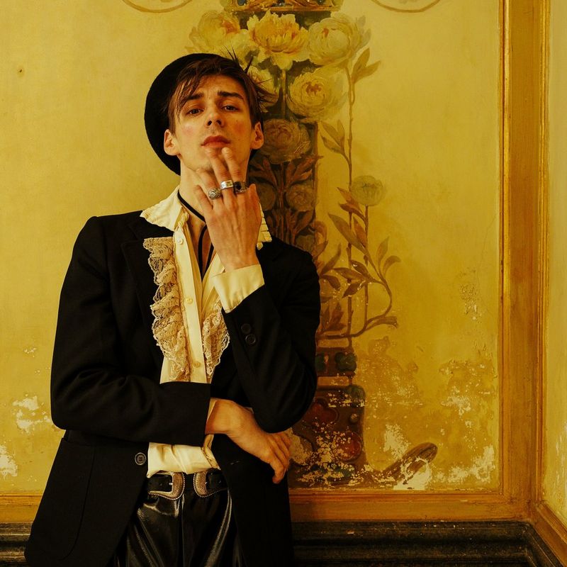 A person wearing a black jacket, a ruffled cream shirt and a black hat leans back against an elaborately painted wall, one hand up to their face with fingers resting on their chin. 