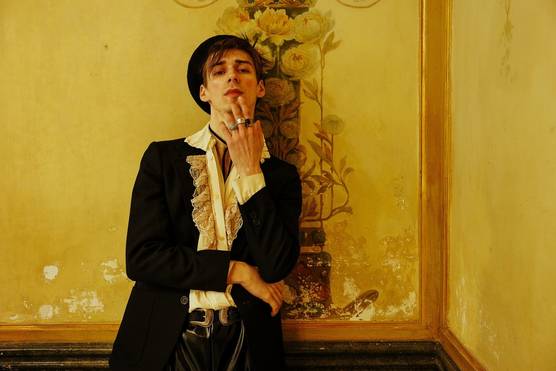 A person wearing a black jacket, a ruffled cream shirt and a black hat leans back against an elaborately painted wall, one hand up to their face with fingers resting on their chin. 