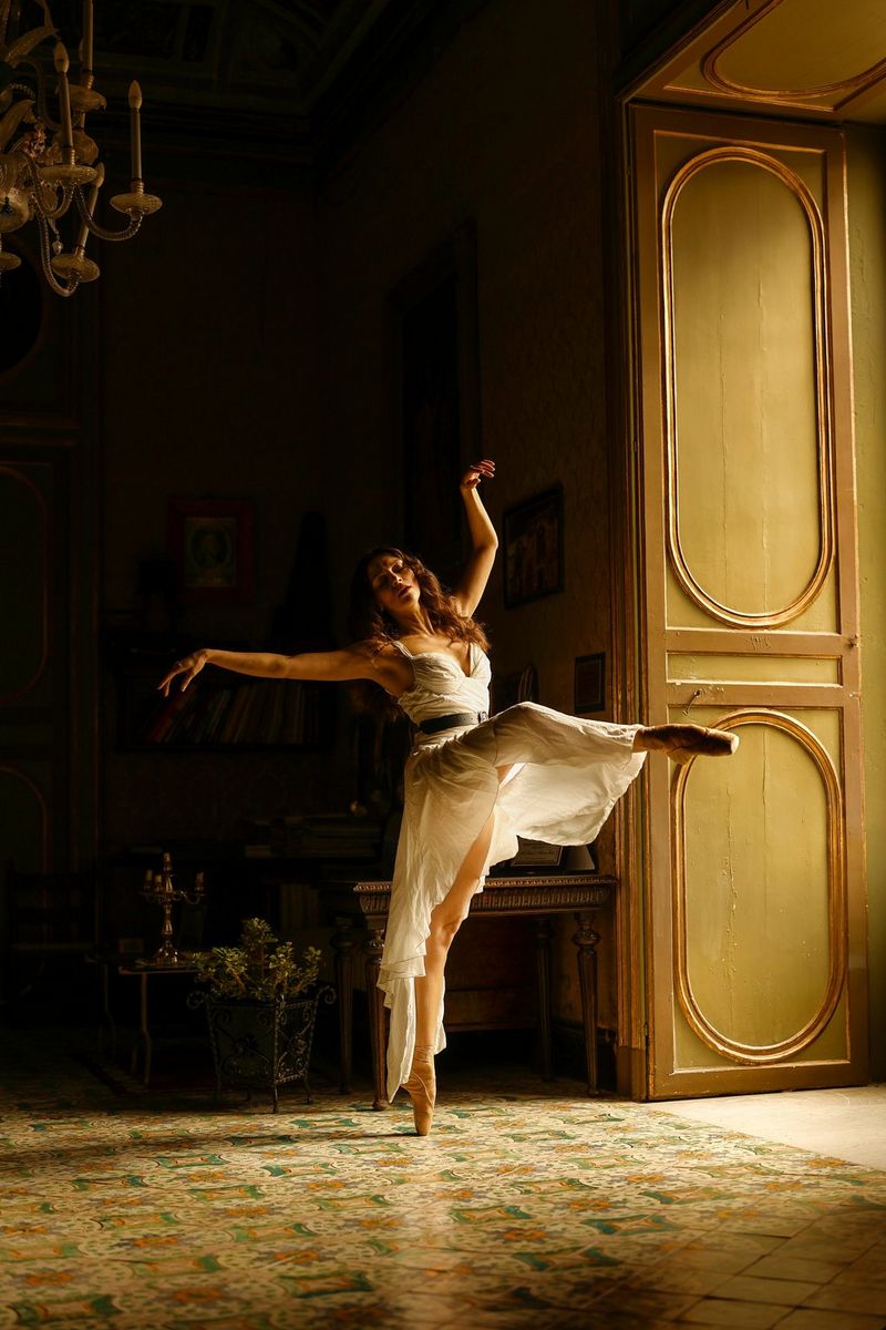 A ballerina dances in the light from an open door. The rest of the room is in shadow.