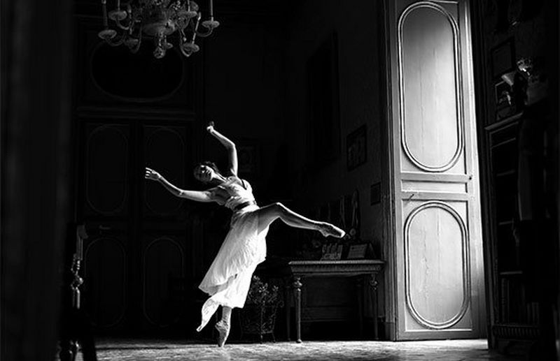 A black and white image of a ballet dancer in motion in a large, opulent room. Sunlight streams through an open door. 