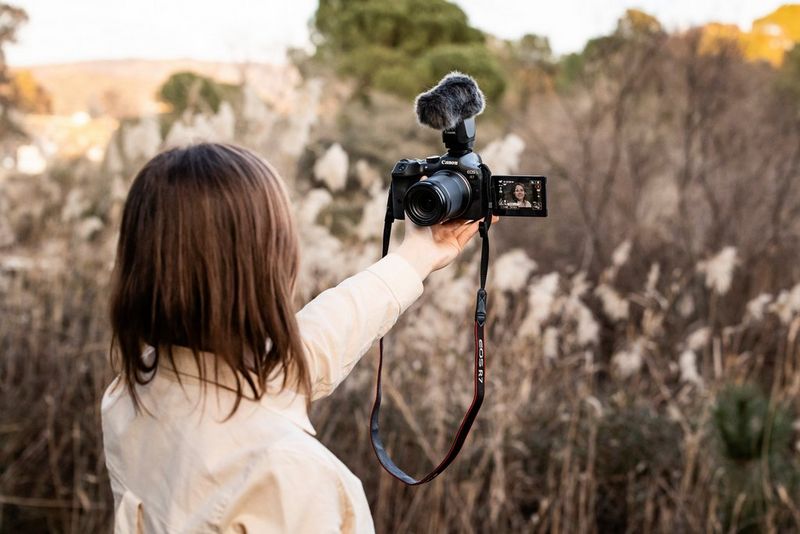 A person in a field holds up a Canon EOS R7 camera pointed at their face.